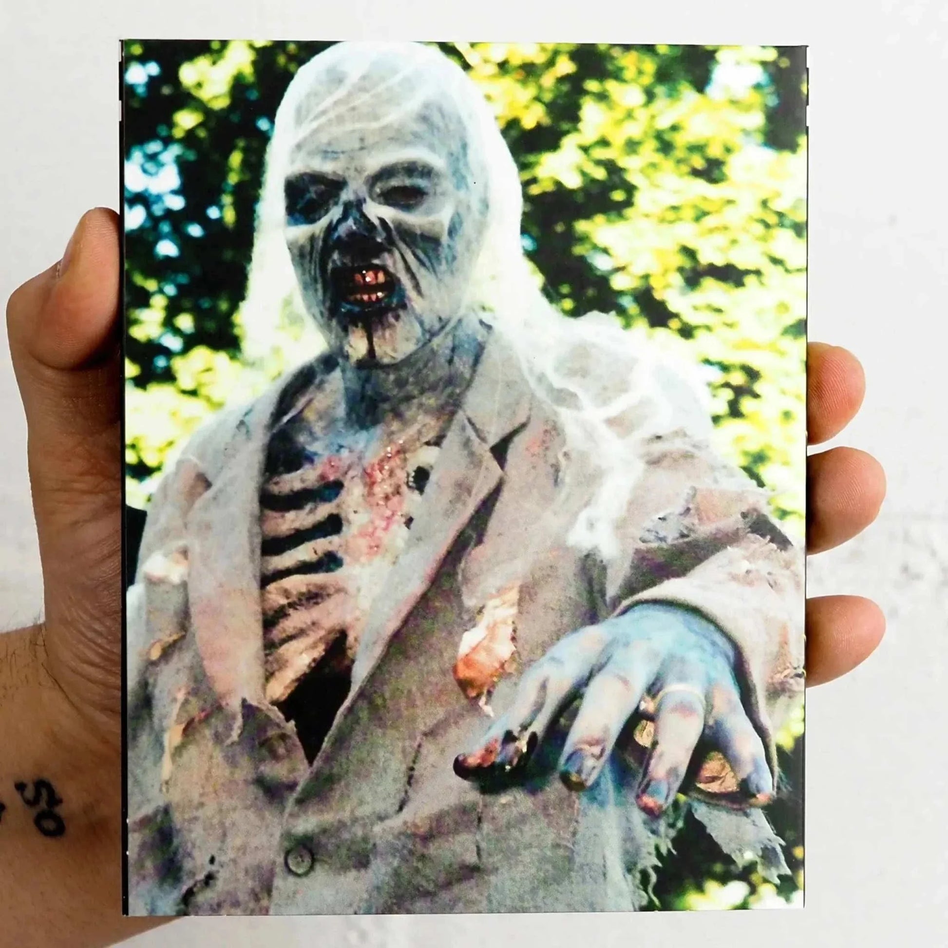 Hand holding Saturn's Core Bone Sickness Blu-Ray slipcover featuring a zombie in tattered suit reaching forward outdoors with green foliage background.